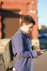 A close up of cute student boy with backpack holds a mobile phone and bottle of water outdoor.