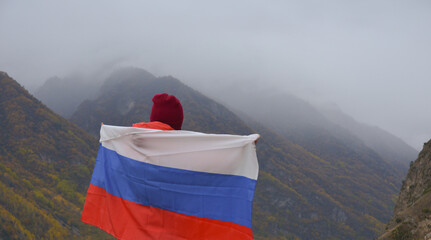 The flag of Russia flutters beautifully behind a woman's back against the backdrop of mountains on a cloudy day. Close-up rear view. Russian Independence Day