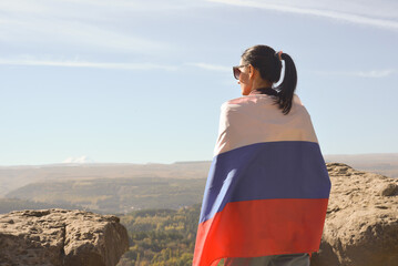 A girl tourist with a Russian flag on her shoulders stands on the top of a mountain and enjoys the...