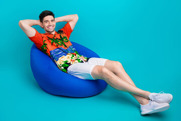 Full size photo of relaxed guy wear tropical shirt stylish shorts sit on bean bag arms behind head...