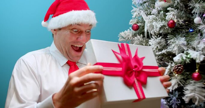 Happy face man delighted with surprise opens box with gifts. Happy smiling dad in Christmas hat Santa Claus receives a gift, rubs his hands before opening box with a gift, looks into box with surprise