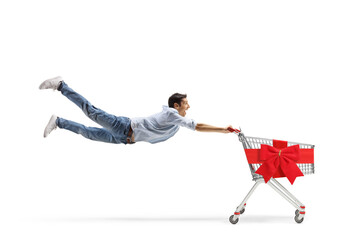 Full length shot of a casual young man flying and holding an empty shopping cart with a red ribbon...