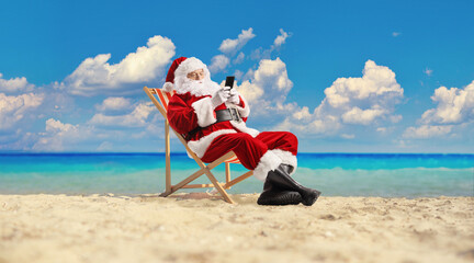 Santa claus sitting at the beach and typing on a smartphone mobile phone