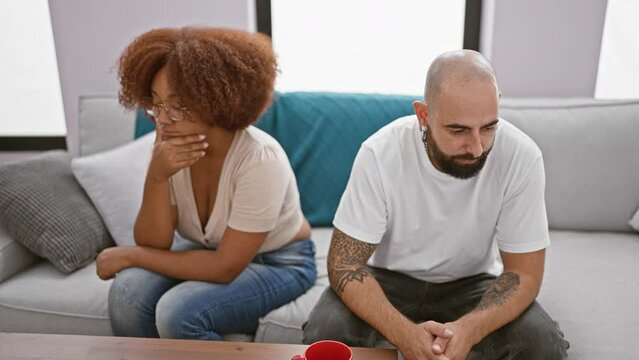 Beautiful interracial couple sitting in silence on living room sofa, stress and anger visible in expression, undercurrent of disagreement at home signifies relationship troubles