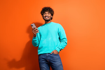 Photo of young happy positive customer in online using smartphone wear stylish jumper indian guy isolated on orange color background