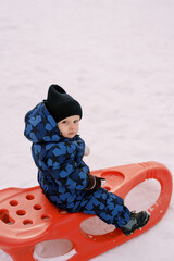 Little boy in overalls sits on a sledge and looks away