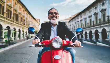 Foto op Plexiglas Excited senior man riding red scooter in Italy, cheerful retired bearded hipster enjoying holiday, motorcycle road trip, trendy vacation lifestyle © Marko