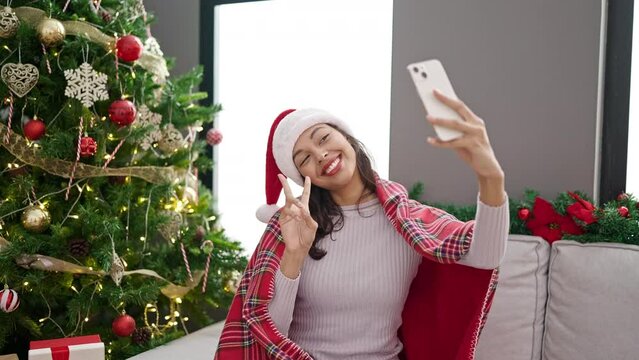 Young beautiful hispanic woman taking selfie by smartphone sitting by christmas tree at home