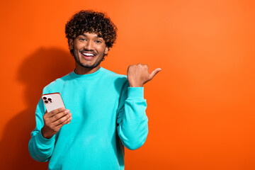 Portrait of arabian young guy direct finger empty space with smartphone indicate new ios features isolated on orange color background