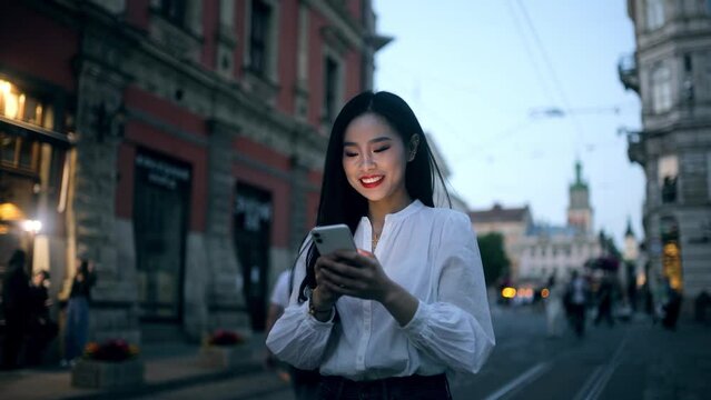 Portrait of young asian woman hold smartphone scrolling social media texting browsing online at urban city Pretty female relax enjoying great late evening while walking down the street outdoors