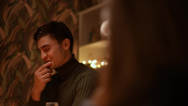 Low-angle view tracking shot of handsome young man talking to friends sitting at festive Christmas dinner table during holiday party in dark room with beautiful lighting. Shooting in slow motion.