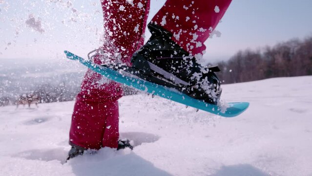 LENS FLARE, SLOW MOTION, CLOSE UP: Lady is running with snowshoes on fresh snow on a beautiful sunny winter day. White snowflakes spray and sparkle in sunlight as she lifts her feet while snowshoeing.