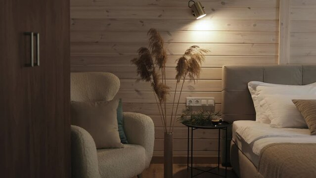 Cozy bedroom with soft chair, bed, warm lamp light on the wooden wall. Pampas grass in tall vase as a interior decor, cozy room in tiny house camera moving forward.