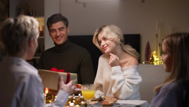 Portrait of happy young woman receiving gift from loving mother-in-law sitting with family at dinner feast table at home on Christmas Eve. Blonde female giving present to beloved daughter on holidays.