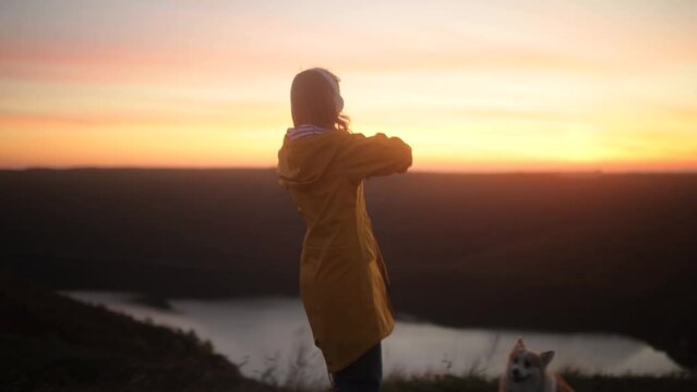 Camera follows young woman in yellow jacket running up on top of mountain summit at sunset, jumps on top of rocks, raises arms into air, happy and drunk on life with her cute fluffy corgi