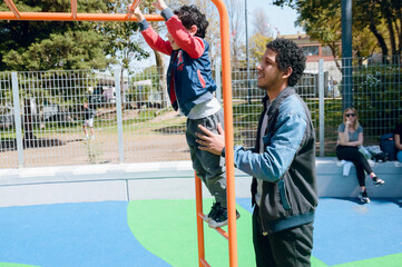 latin young father helping his son playing in public park games - obrazy, fototapety, plakaty