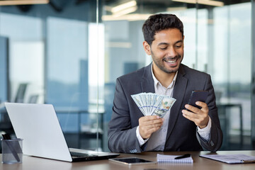 Young successful businessman inside office workplace holding phone and money cash US dollars in...