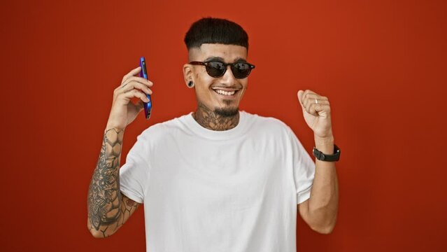 Cheerful young latino man dancing with joy while listening to catchy tunes on his phone, isolated over a vibrant red background