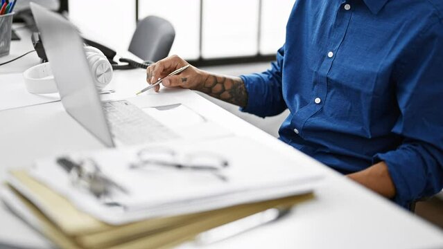 Young chap with latin roots excelling in his business work, sitting at his office table, tattooed yet purely professional, personifying success in the modern workplace.