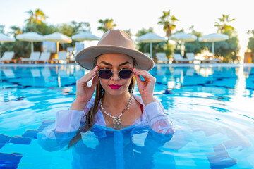 Stylish woman with sunglasses in pool, white shirt contrasting with blue water