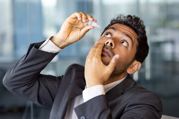Businessman has severe eye pain, drips drops for pain relief, man at workplace inside office,...