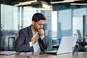 Sick businessman coughing inside office at workplace, man in business suit working with laptop,...