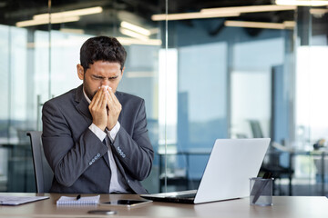 A man sneezes into a tissue at a workplace inside an office, a businessman is sick with a runny nose, works at a workplace with allergies, uses a laptop at work. - obrazy, fototapety, plakaty