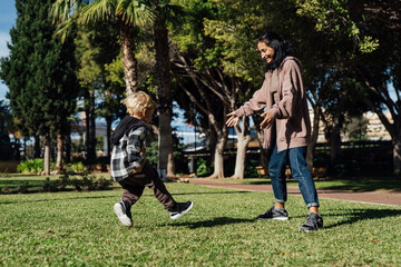 Fototapeta premium Mother and her three-year-old son run on the grass in the park, playing, hugging, smiling, and giving each other kisses