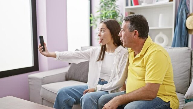 Joyous hispanic father and daughter taking smiling selfie picture on smartphone together, sitting on sofa in warm family home living room