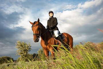 Horsewoman in equestrian sports gear, riding a horse, against an expressive sky, horseback riding in the open air - 676570531