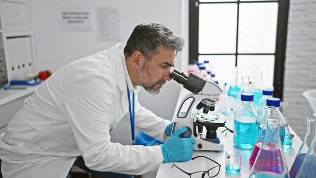 Confident young hispanic scientist with grey hair, smiling as he works with a microscope in the lab, making medical discoveries