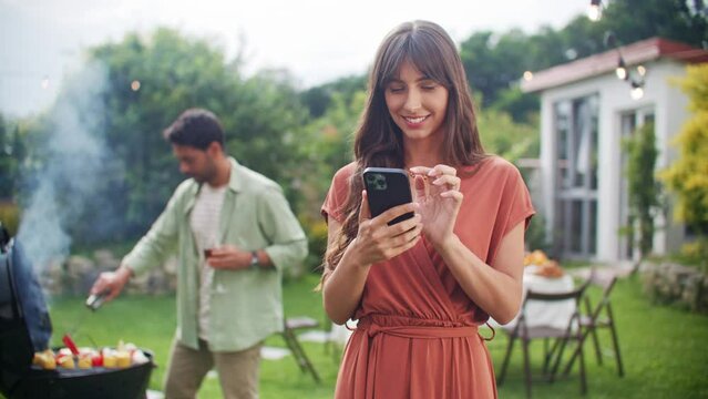 Portrait of young beautiful woman holding smartphone, smiling, texting, chatting and looking at phone screen. Leisure, holidays, eating, people and food concept - friends having party at house yard.