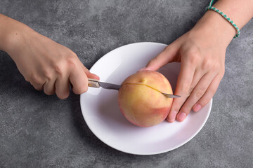 Authentic female hands cutting a fresh ripe peach on white plate on a grey table background, close up. Healthy summer snack, food lifestyle