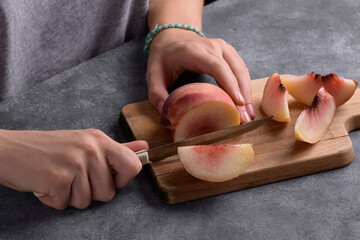 Authentic female hands cutting a fresh ripe peach on wooden cutting board on a grey table...