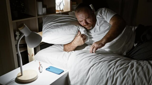 Handsome middle-aged caucasian man, comfortably resting in bed, sleepily waking up to the morning light. looking at his smartphone to check the time before standing up in his bedroom.