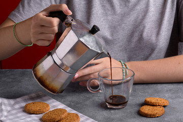 Young woman pouring espresso coffee from moka pot coffee maker into cup in kitchen table at home