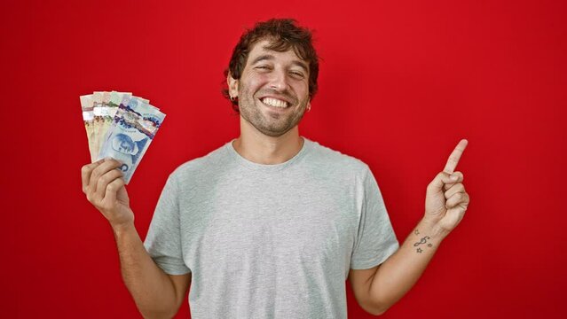 Hopeful young man joyfully holding canadian dollar banknotes, confidently pointing to the side with a friendly smile, isolated on a red background.
