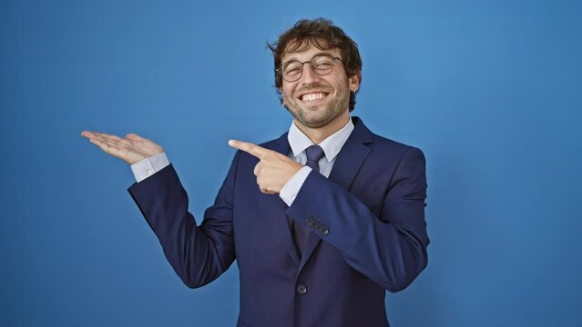 Cheerful young man in a business suit, amazed while confidently presenting and pointing with a smile on isolated blue background