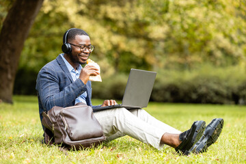 Man in business suit sitting on grass, eating his lunch and surfing the internet in city park.
