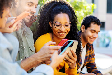 Millennial group of students using mobile phones together at campus college. African american woman having fun watching social media content on cellphone app sitting with her friends outdoors.
