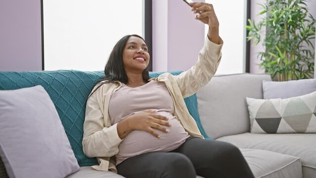 Joyful young pregnant woman, sitting and chatting on the phone, takes a selfie of her maternity glow, touching her belly with happiness in the comfort of her living room
