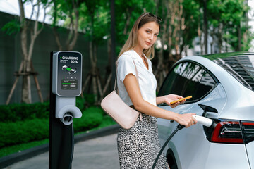 Young woman travel with EV electric car charging in green sustainable city outdoor garden in...