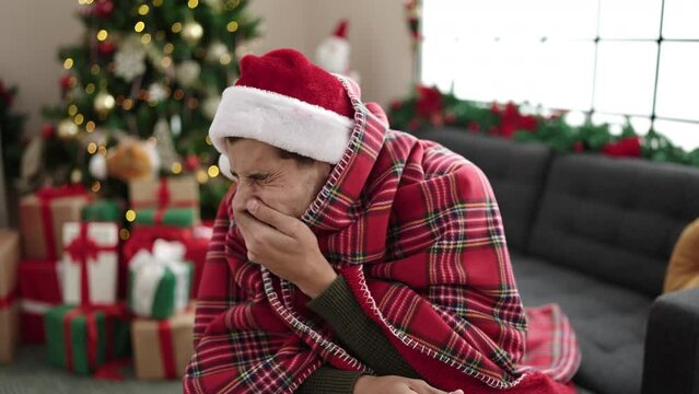 Young hispanic man feeling cold covered with blanket sitting on sofa by christmas tree at home
