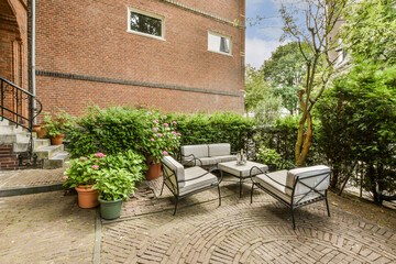 a patio with chairs and plants on the ground in front of a red brick building, surrounded by green...