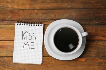 Notebook with phrase Kiss Me and cup of coffee on wooden table, flat lay