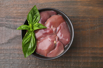 Bowl of raw chicken liver with basil on wooden table, top view