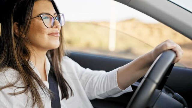 Young beautiful hispanic woman driving a car smiling wearing glasses on the road