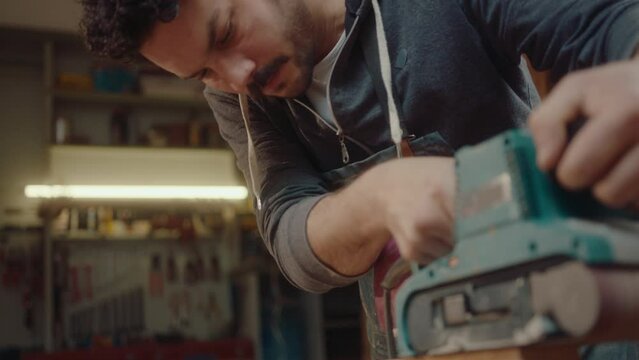 Young woodworker in apron polishing wooden board with belt sander during workday in the woodshop. Tilt-up shot