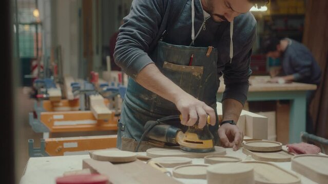 Young male artisan finishing wooden serving trays with random orbit sander and sandpaper, creating handmade home decor in the woodshop. Tilt-up shot