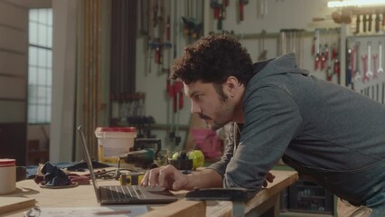 Young male woodworker leaning on workbench and using the Internet on laptop during the day in carpentry workshop. Side view, medium shot